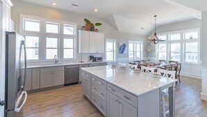 A spacious kitchen with a large island, white cabinetry, and ample natural light from large windows. - Sun Realty gorgeous Kitty Hawk home with elevator, just steps from the beach (Kitty Hawk)