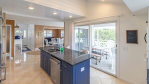 A modern kitchen with a dark blue island, white cabinetry, and a large window leading to an outdoor patio. - Sun Realty warm Duck NC vacation rental, just a short walk to the beach (Duck)