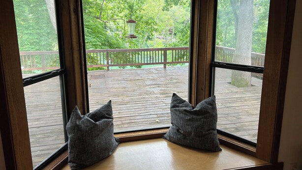 A corner window with two gray pillows on a wooden ledge, overlooking a wooden deck with a railing and trees.