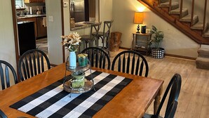 A dining area with a wooden table, black and white checkered table runner, and black chairs. There is a kitchen visible through an open door, a staircase with wooden railings, and a ceiling fan.