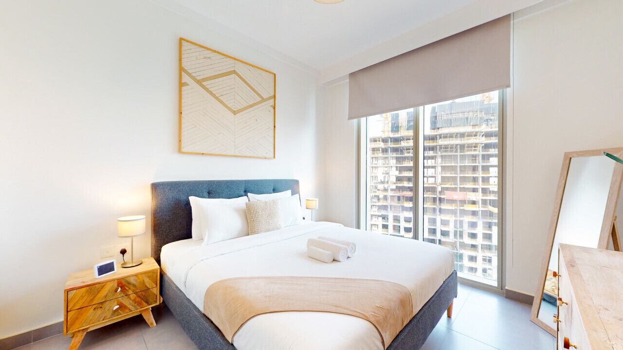 A modern hotel room with a large bed, a nightstand with a lamp, and a view of a cityscape through the window.