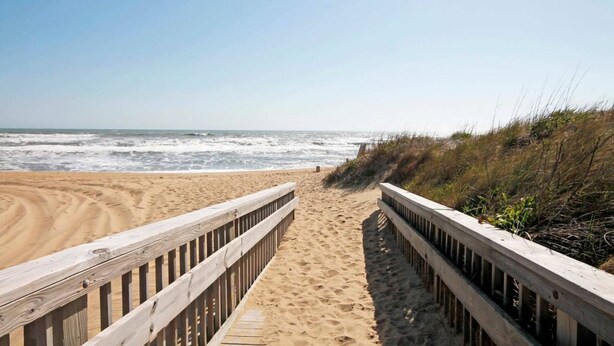 Une promenade en bois mène à une plage de sable, avec l’océan et ses vagues en arrière-plan.