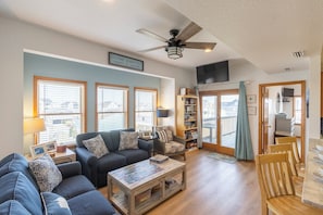 A living room with a sofa, coffee table, and a ceiling fan. There are windows with blinds, a bookshelf, and a television.
