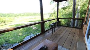 Une terrasse en bois offrant une vue sur une rivière et une forêt. On y trouve trois tabourets et un banc.
