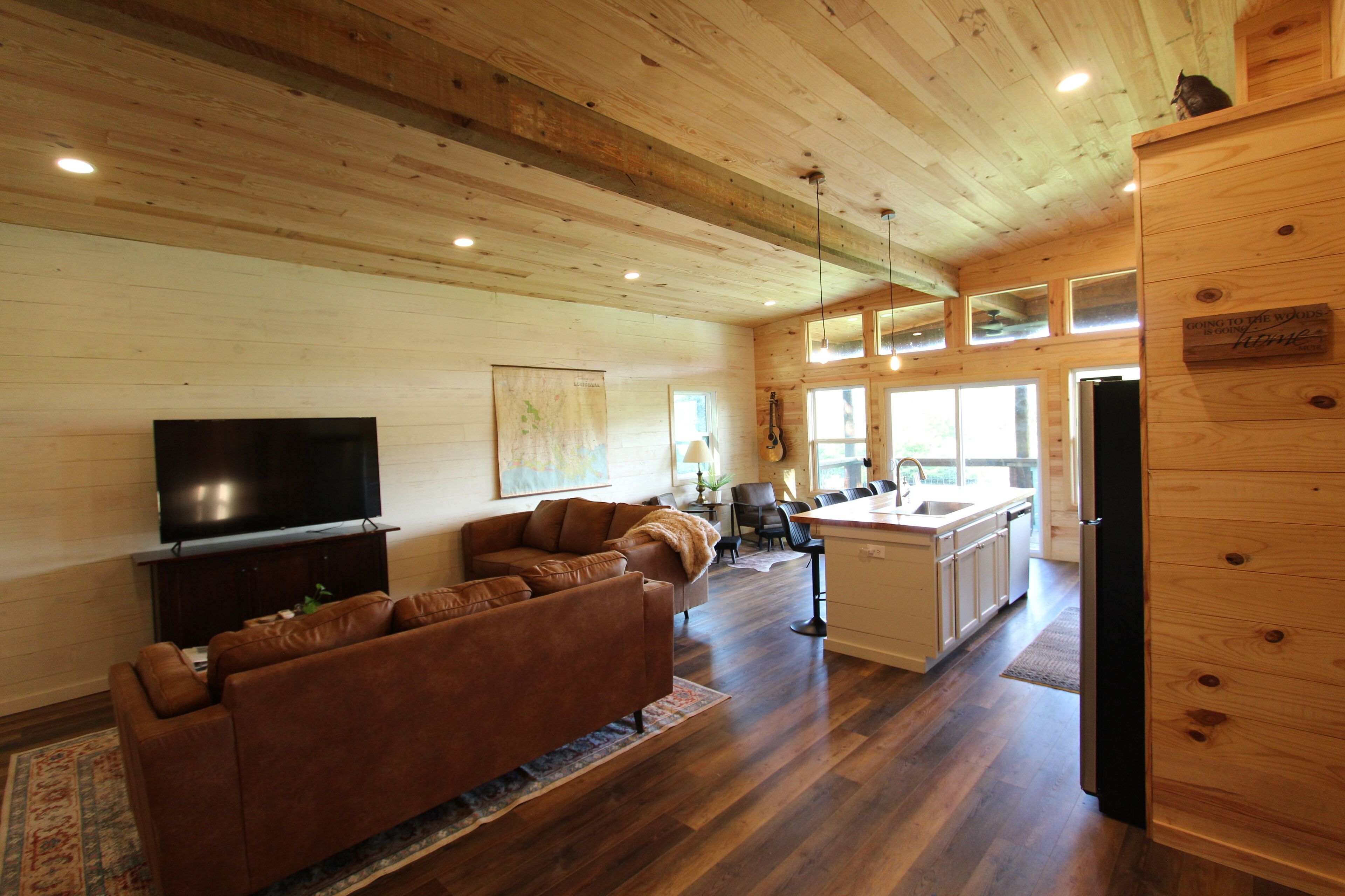Una sala de estar moderna con un sofá de cuero marrón, un televisor de pantalla plana, pisos de madera y una zona de cocina con armarios de madera y un refrigerador.