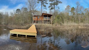 Une cabane en bois dotée d’une terrasse, entourée d’une forêt et bordée par un plan d’eau.
