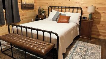 Un dormitorio con estructura de cama de metal, un banco, cabecera de madera, mesita de noche, lámpara y un reloj de pared.