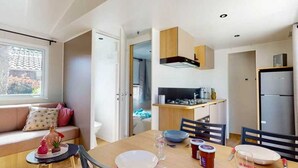 Une cuisine compacte équipée d’une cuisinière, d’un évier et d’un réfrigérateur. On y trouve une table à manger avec des chaises et une fenêtre avec des stores.