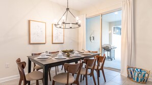 A dining area with a large table set for six, surrounded by wooden chairs. There are framed artworks on the wall, a chandelier above the table, and a wicker basket with cushions to the side. - Beautiful Storey Lake Retreat Minutes From Disney (Kissimmee)