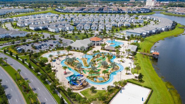 An aerial view of a residential area with a central swimming pool complex, surrounded by single-family homes, and a road curving around the property. - Beautiful Storey Lake Retreat Minutes From Disney (Kissimmee)