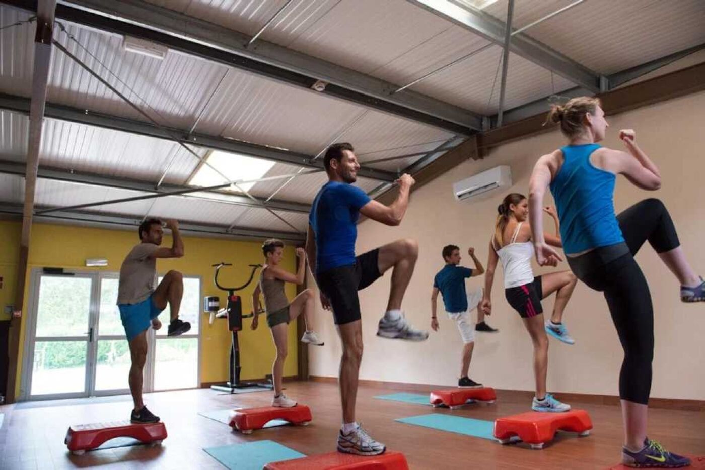 Un groupe de personnes fait de l’exercice sur des plateformes d’aérobic dans une salle de sport équipée de vélos d’exercice et dotée d’une grande fenêtre.