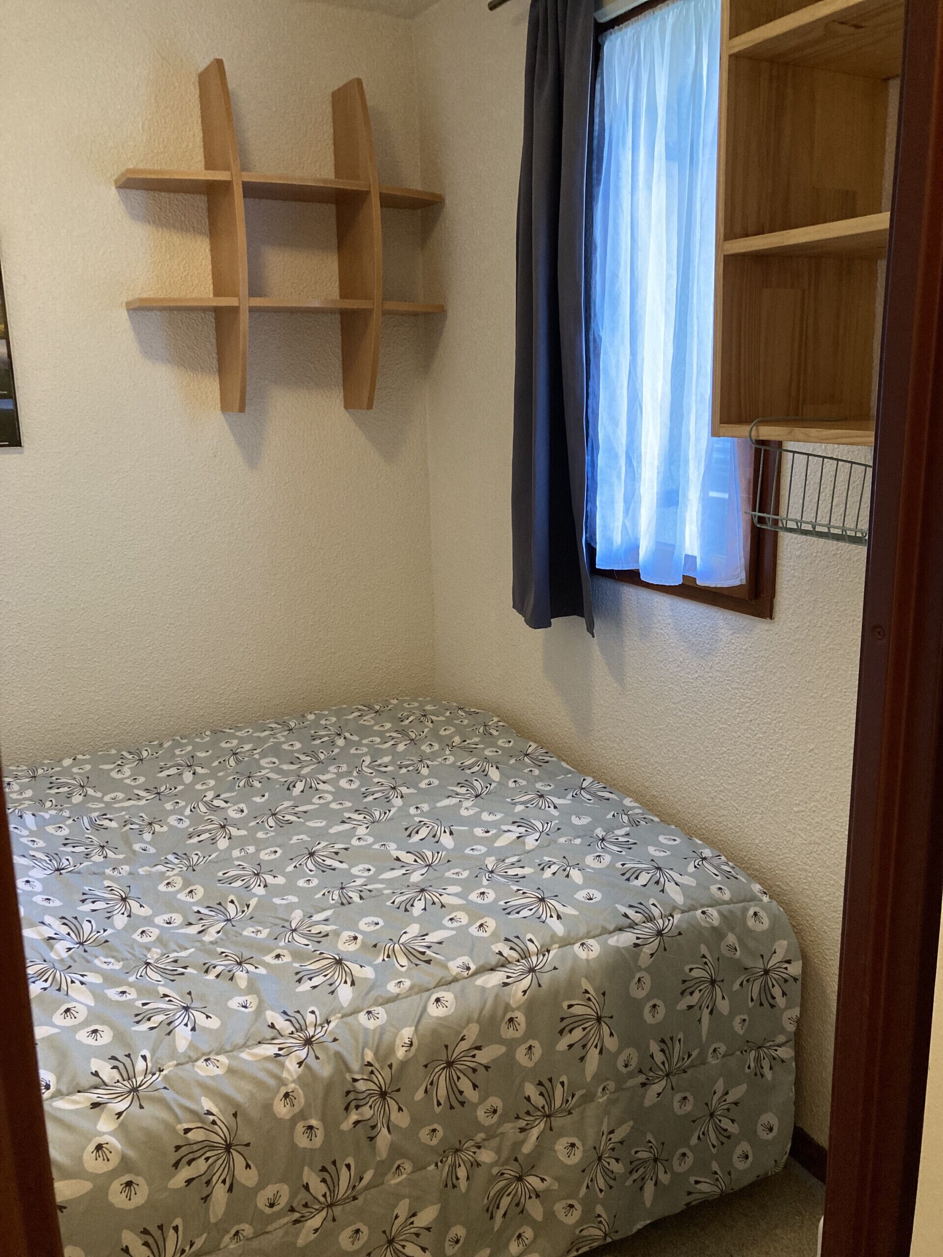 A small, single-bed room with a floral-patterned bedspread, wooden shelves, and a window with a curtain.