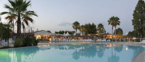 A swimming pool with lounge chairs and umbrellas, surrounded by palm trees and buildings.