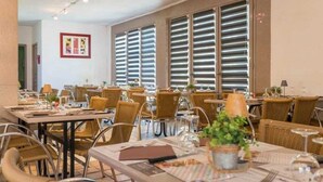 En restaurant med fletstole og borde, store vinduer med rullegardiner og et billede i ramme på væggen.