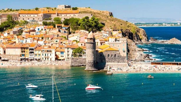 A coastal town with historic buildings, a prominent tower, and a beach with people and boats.