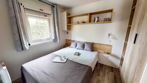 A neatly arranged bedroom with a bed, bedside table, and a shelf above the bed. - ZEcamping 4* familial 100m plage toboggans locatif clim 28m2 8 pers (Canet-en-Roussillon)