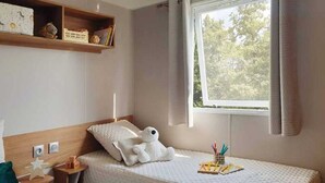 A neatly arranged bedroom with a bed, a nightstand, a shelf with books and decorative items, and a window with a view of trees. - ZEcamping 4* 1km plage toboggans pet friendly animations locatif 36m2 (Les Mathes)