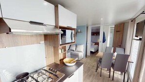 A compact kitchen with a stove, a bowl on the counter, and a dining area with chairs and a table. - ZEcamping 4* 1km plage toboggans dog friendly locatif clim 36m2 8 pers (Les Mathes)