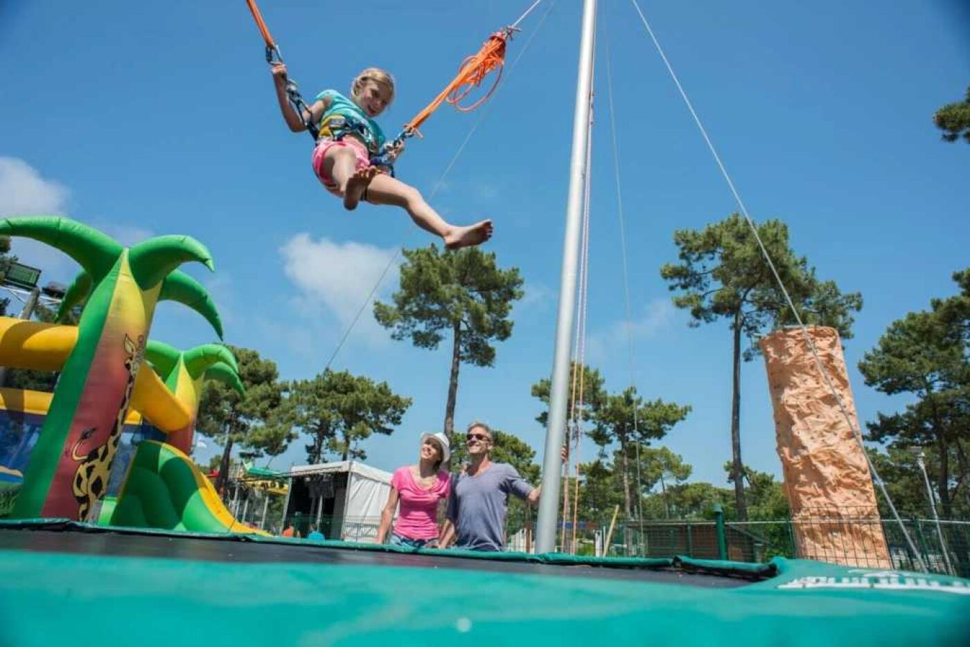 Une personne sur une balançoire, avec un toboggan gonflable vert et un grand arbre en arrière-plan.