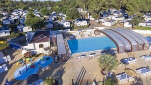 Ein großer Swimmingpool mit überdachtem Bereich, umgeben von Liegestühlen und Sonnenschirmen, inmitten eines Campingplatzes mit Mobilheimen im Hintergrund.