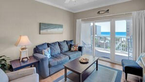 Ein Wohnzimmer mit einer Couch, einem Couchtisch und einer Lampe. An der Wand hängt ein Gemälde, und durch die Balkontüren ist der Blick auf den Ozean.