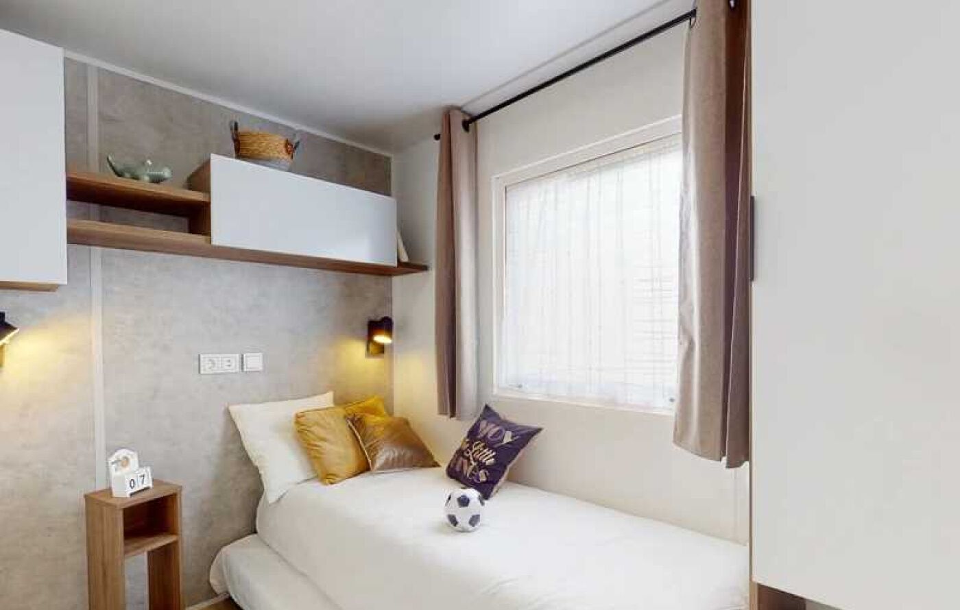 Ein kleiner Raum mit einem Bett, einem Regal mit Dekoration, einem Fenster mit Vorhängen und einer Wandleuchte.