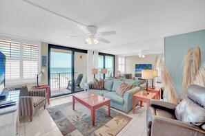 Een ruime woonkamer met een grote bank, een salontafel en een televisie. Er is een balkon met uitzicht op zee.