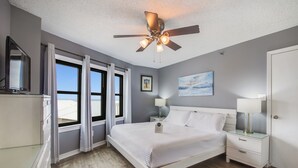 Un dormitorio con cama, ventilador de techo, mesita de noche, lámpara, cómoda y ventana con cortinas.
