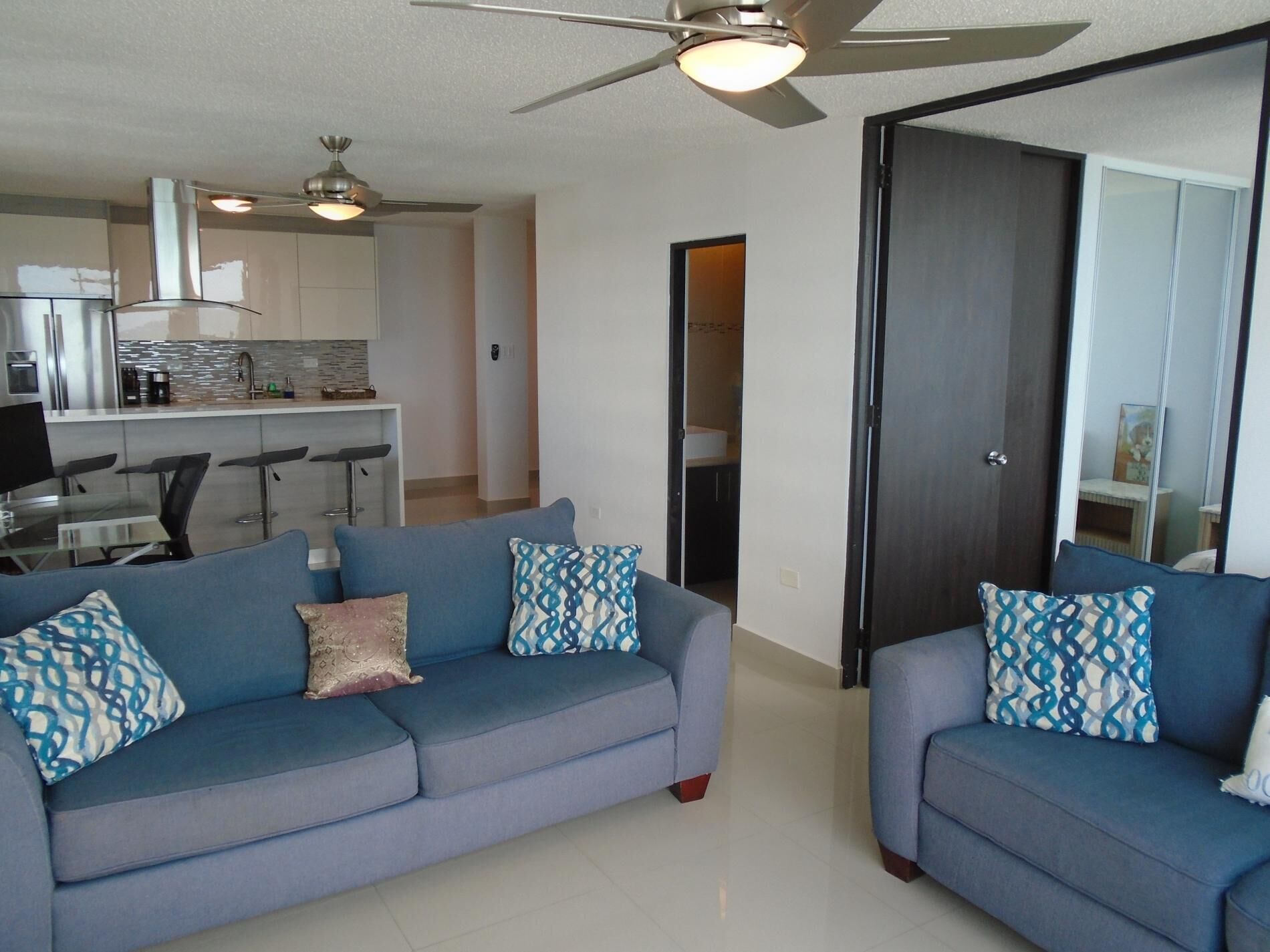 A modern living room with two sofas, a ceiling fan, and a kitchen area in the background.
