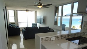 A modern kitchen with a central island, a dining area with a view of the ocean, and a living room with a sofa and a TV. - Lucille's Corner (Carolina)