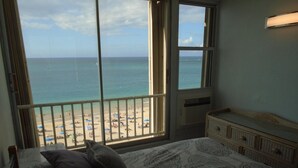 A bedroom with a large window overlooking a beach, a bed with patterned bedding, and a dresser with drawers. - Lucille's Corner (Carolina)