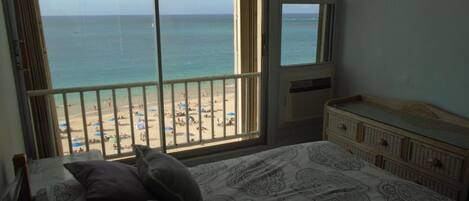 A bedroom with a large window overlooking a beach, a bed with patterned bedding, and a dresser with drawers.