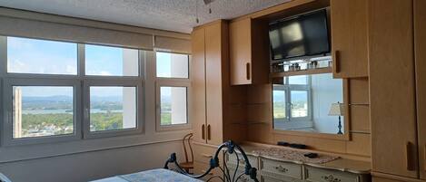 A bedroom with a bed, a ceiling fan, a TV, wooden cabinets, and a window with a view of a landscape.