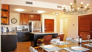 A modern kitchen with wooden cabinets, a refrigerator, a microwave, and a coffee maker. There is a dining area with a table set for two, chairs, and a chandelier. - Blue Horizon (Humacao)
