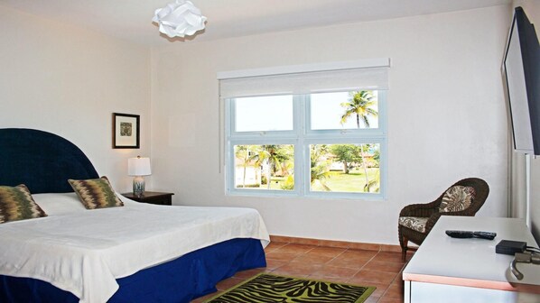 A bedroom with a bed, a chair, a desk, a lamp, and a window with a view of palm trees.
