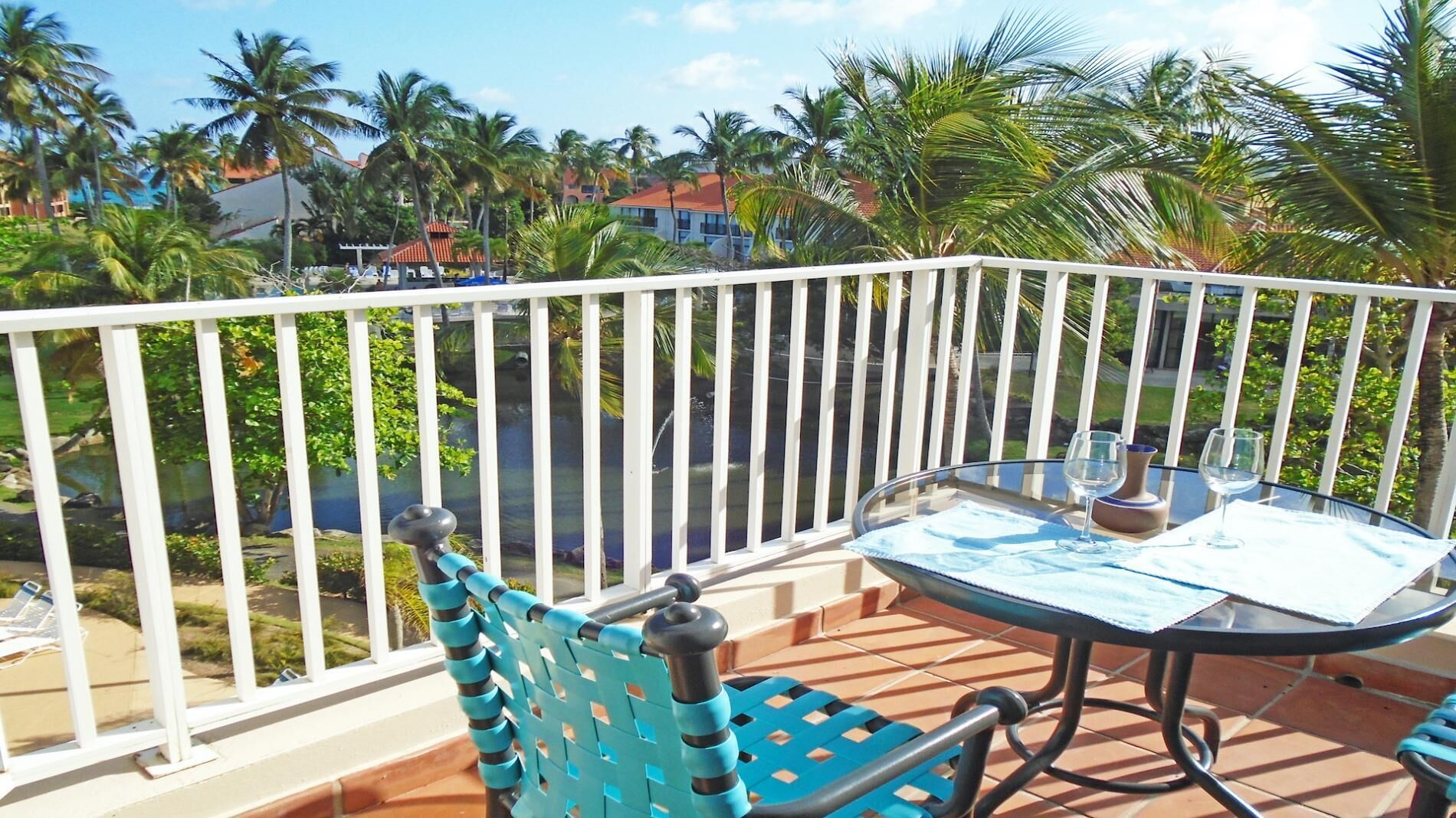 A balcony with a glass table and chairs, overlooking a pool and palm trees.