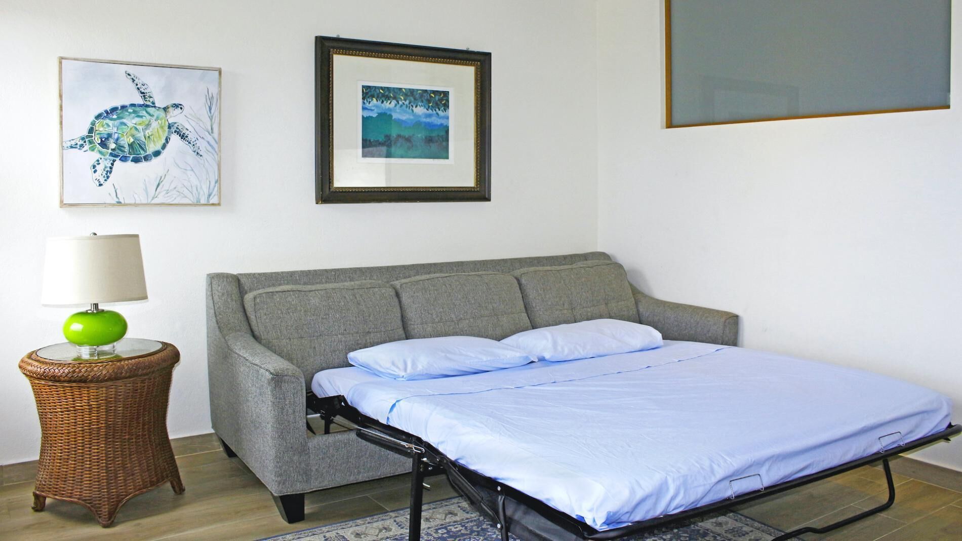 A bedroom with a grey sofa bed, a green lamp, a wicker side table, and two framed pictures on the wall.