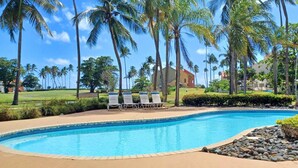 Una piscina con sillones de descanso, rodeada de palmeras y un campo de golf al fondo.