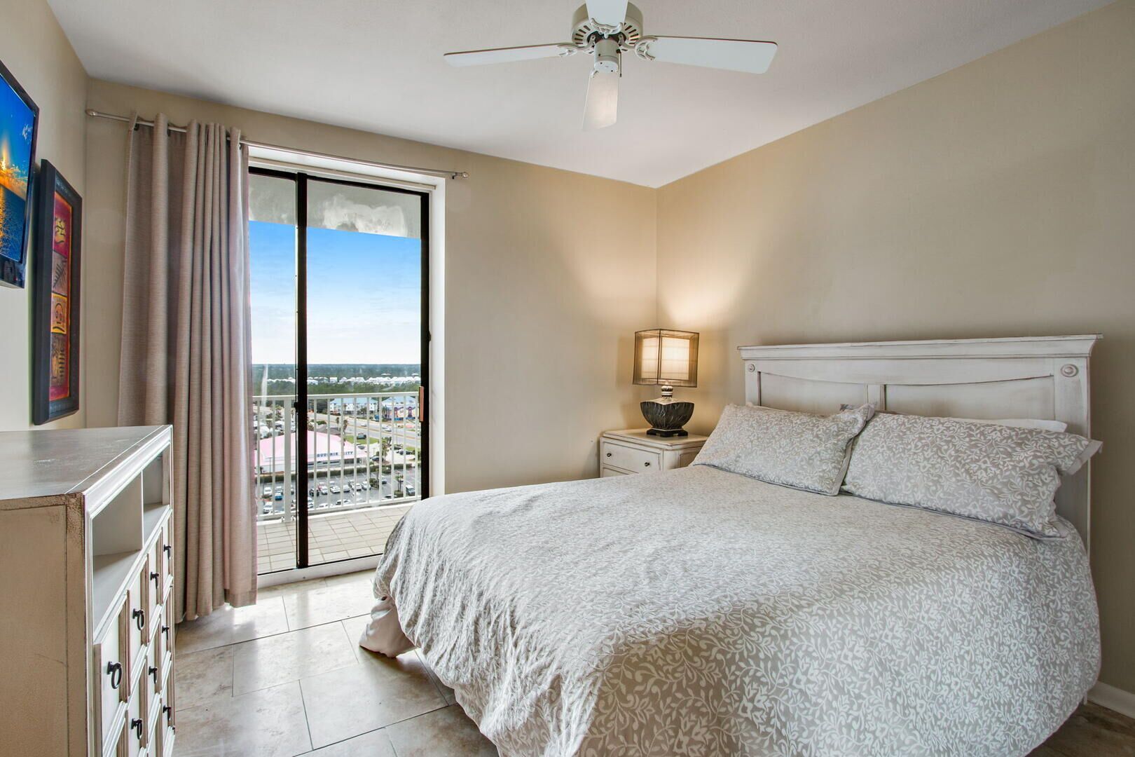 A bedroom with a large bed, a nightstand with a lamp, a ceiling fan, and a view of a parking lot through a sliding door.