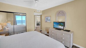 Habitación de hotel con una cama grande, un televisor en una cómoda, un espejo y vista al océano a través de un ventanal corredera.
