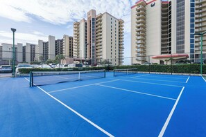 A tennis court with a net in the middle, surrounded by a fence, with tall residential buildings in the background.