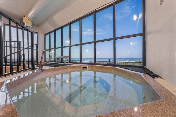 An indoor swimming pool with clear glass walls, a metal railing, and a view of the beach.