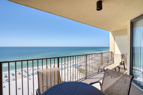 A balcony with a view of a beach and ocean. There are chairs and a table on the balcony.