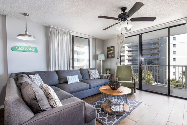 A modern living room with a grey sofa, a round coffee table, a ceiling fan, and a balcony with a view of a residential building.