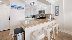 Una cocina con armarios blancos, encimera de granito, microondas y cafetera. Hay tres taburetes blancos y un cubo de basura.