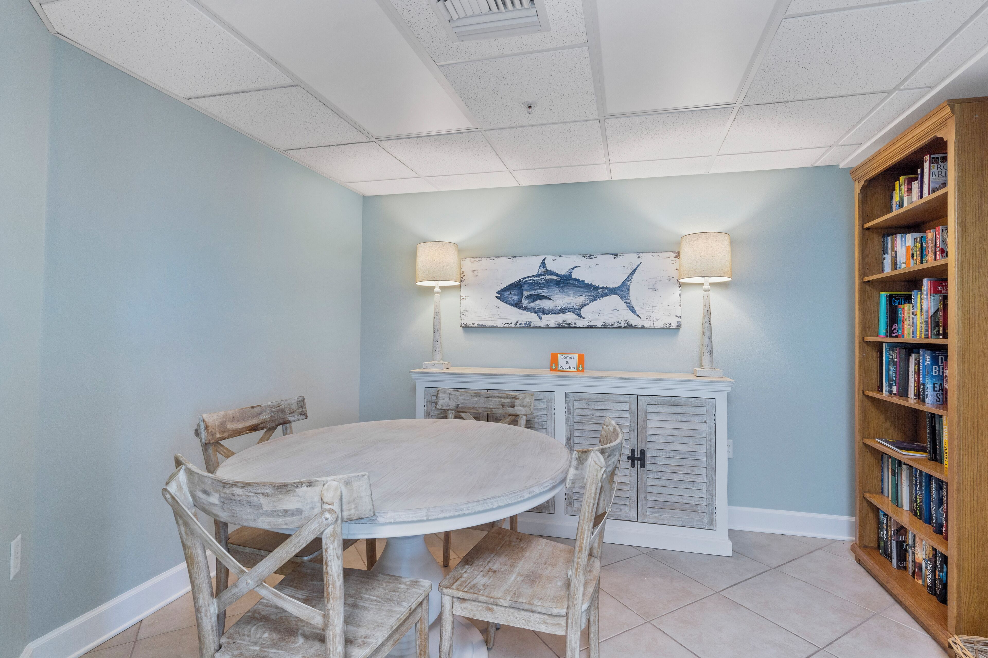 Uma sala de jantar com uma mesa redonda, cadeiras de madeira, um armário branco com uma pintura de peixe e uma estante de livros.