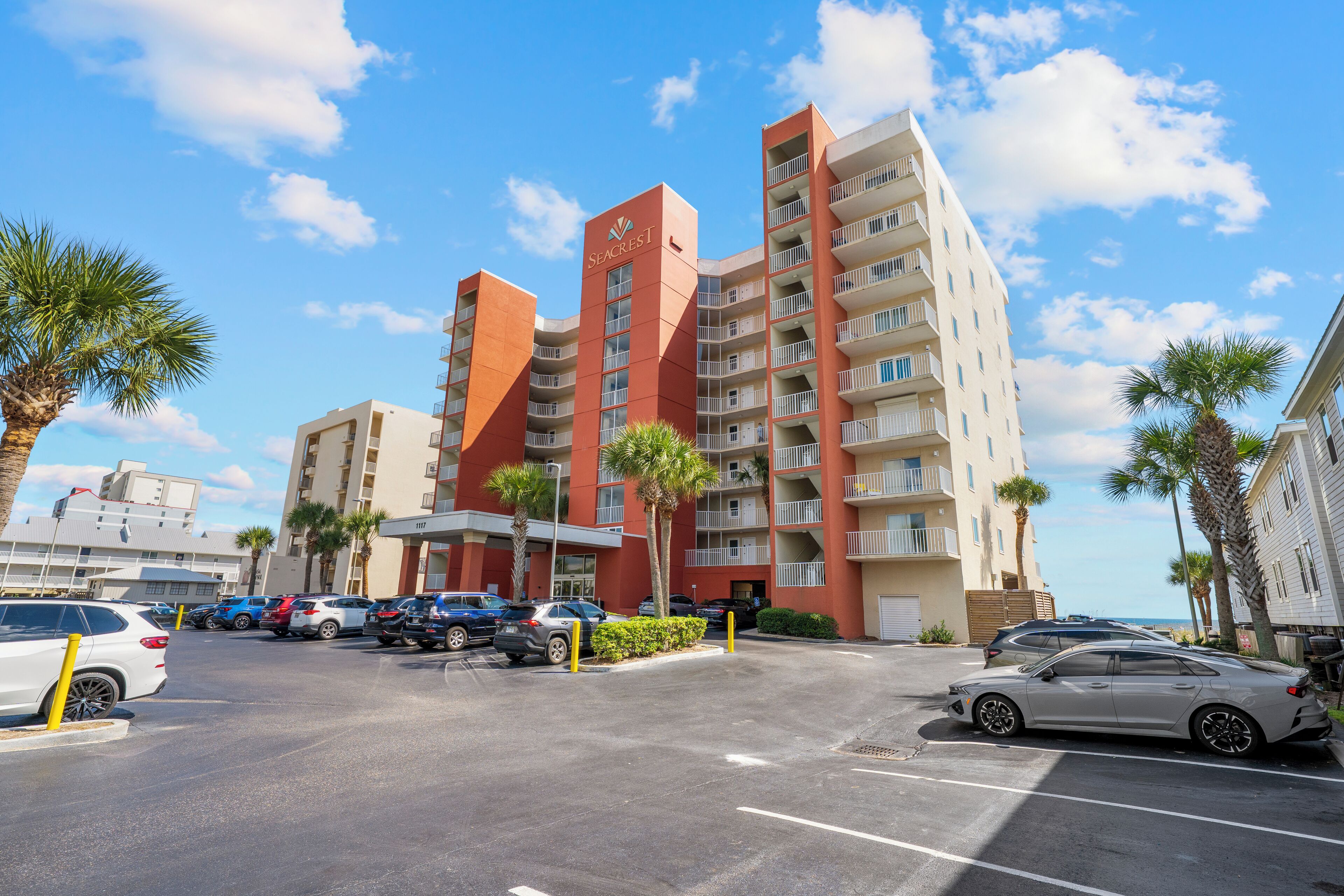 A multi-story hotel with a red and beige facade, a parking lot with several cars, and palm trees in the foreground.