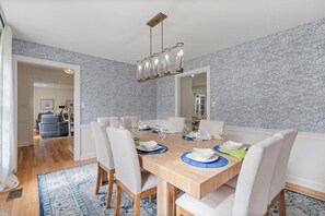 Comedor con mesa de madera dispuesta para cuatro, rodeada de sillas blancas, con papel tapiz estampado y una araña de techo.