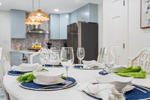 A dining table set with plates, bowls, and glasses, with a modern kitchen in the background. - 4BR, Best Location, Pool, Hot tub, Game Room (raleigh)