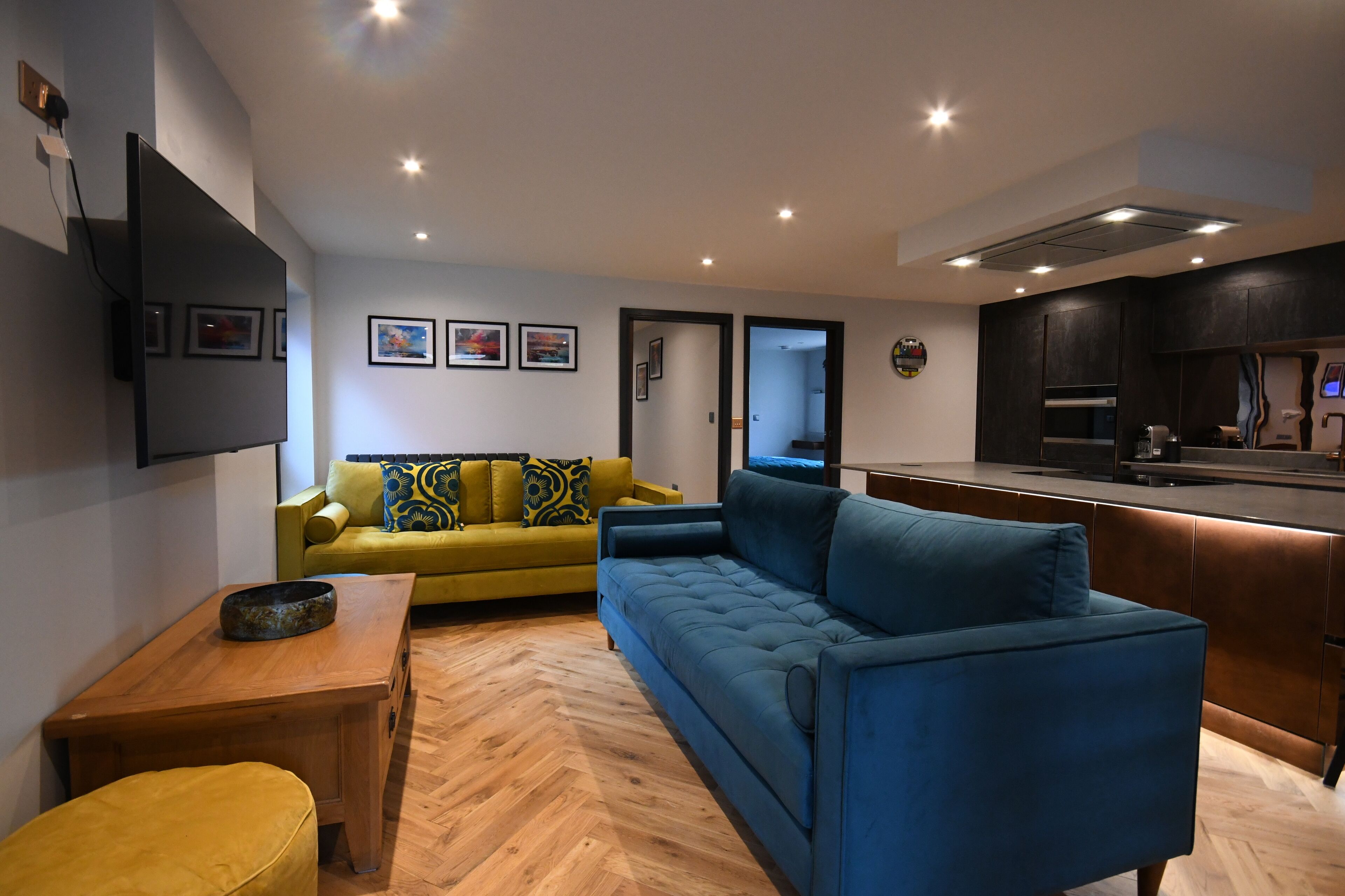 A modern living room with a sofa, a coffee table, and a dining table. There is a kitchen area with a stove and cabinets.